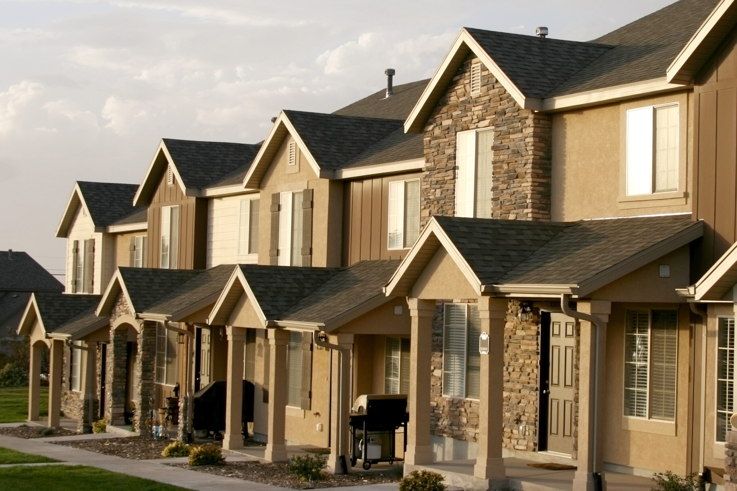 Row of modern two-story townhouses with stone and stucco exteriors, covered front porches, and manicured lawns, photographed in daylight.