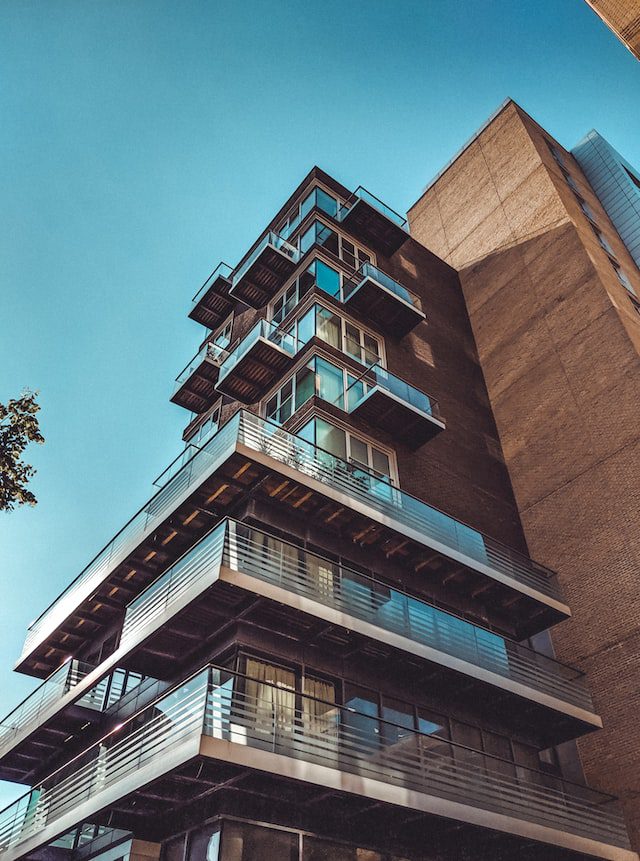 Low-angle view of a modern apartment building with multiple glass balconies and metal railings against a clear blue sky.