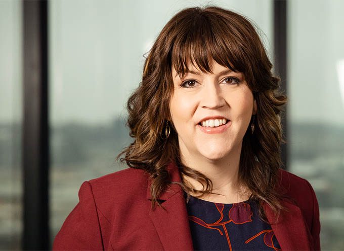 Woman with wavy brown hair wearing a maroon blazer and patterned blouse, smiling, standing in front of large windows with a blurred cityscape background.