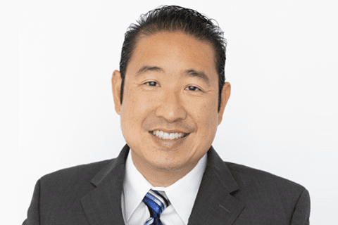 A man wearing a dark suit, white shirt, and striped tie smiles at the camera against a plain white background.