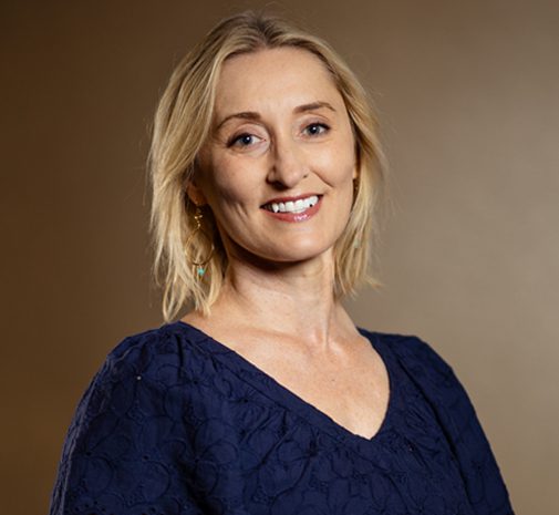 A woman with shoulder-length blonde hair, wearing a navy blue top, smiles at the camera against a plain brown background.