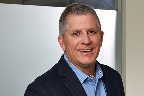 A middle-aged man with short gray hair wearing a navy blazer and light blue shirt, smiling in front of a frosted glass background.