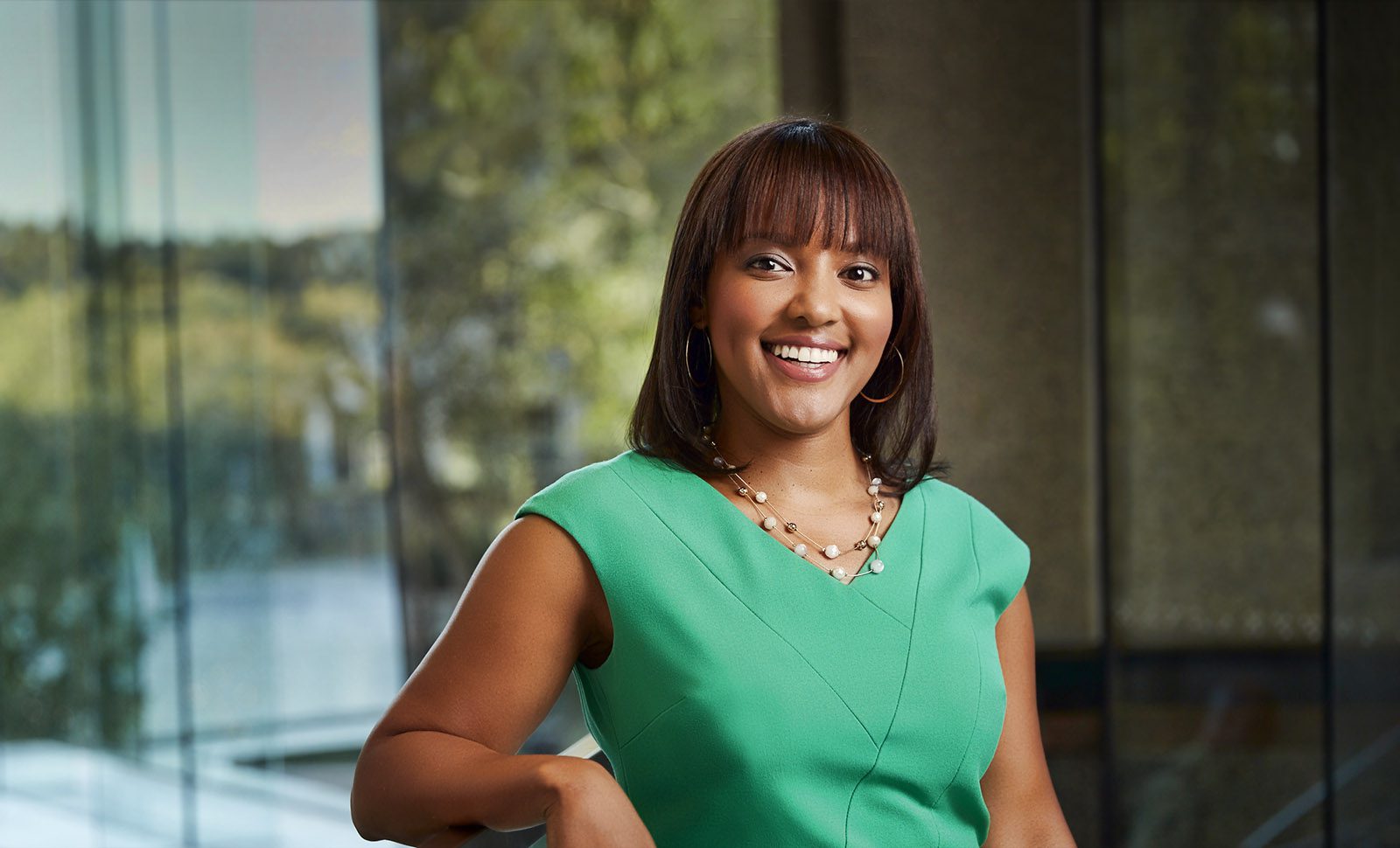 A woman in a green dress stands indoors, smiling at the camera. She has straight shoulder-length hair and is wearing a necklace and hoop earrings. A blurred window background is visible.