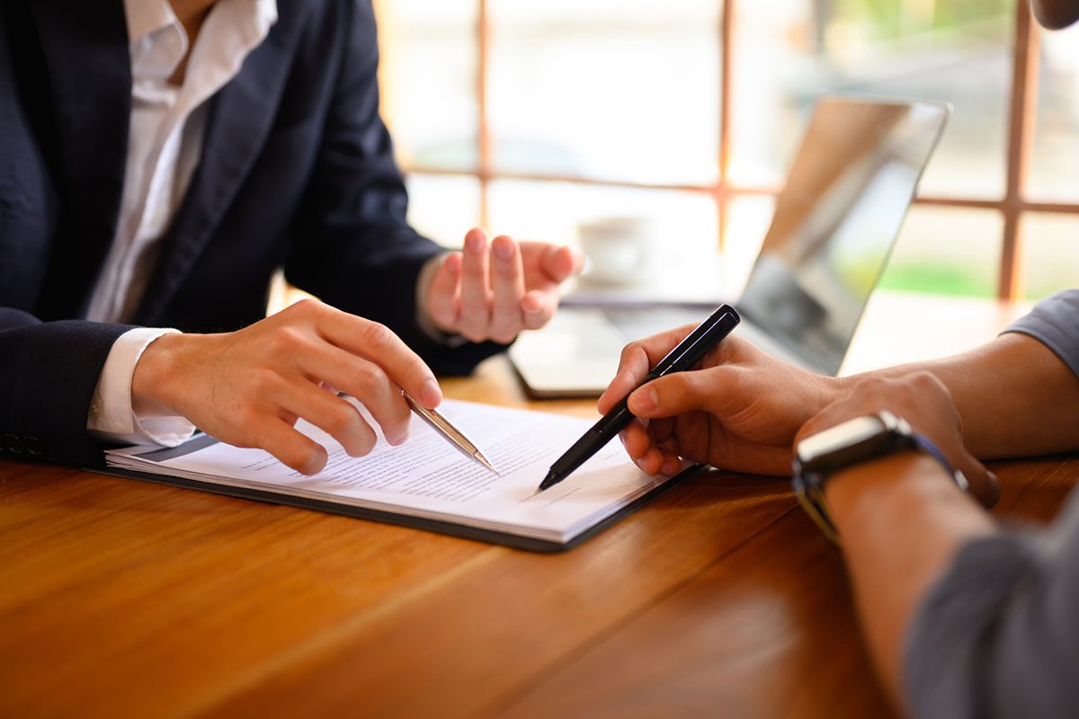 Two people in business attire review and discuss a document on a clipboard at a wooden table, with a laptop in the background.