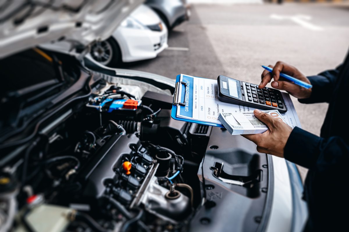 Person standing next to an open car hood, holding a clipboard with documents, a calculator, and a pen, appearing to calculate or review automotive costs or repair estimates.