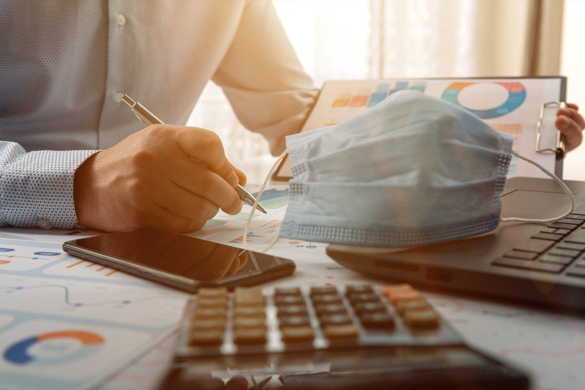 Person working at a desk with charts, a mask, smartphone, laptop, and calculator, holding a pen and reviewing documents.