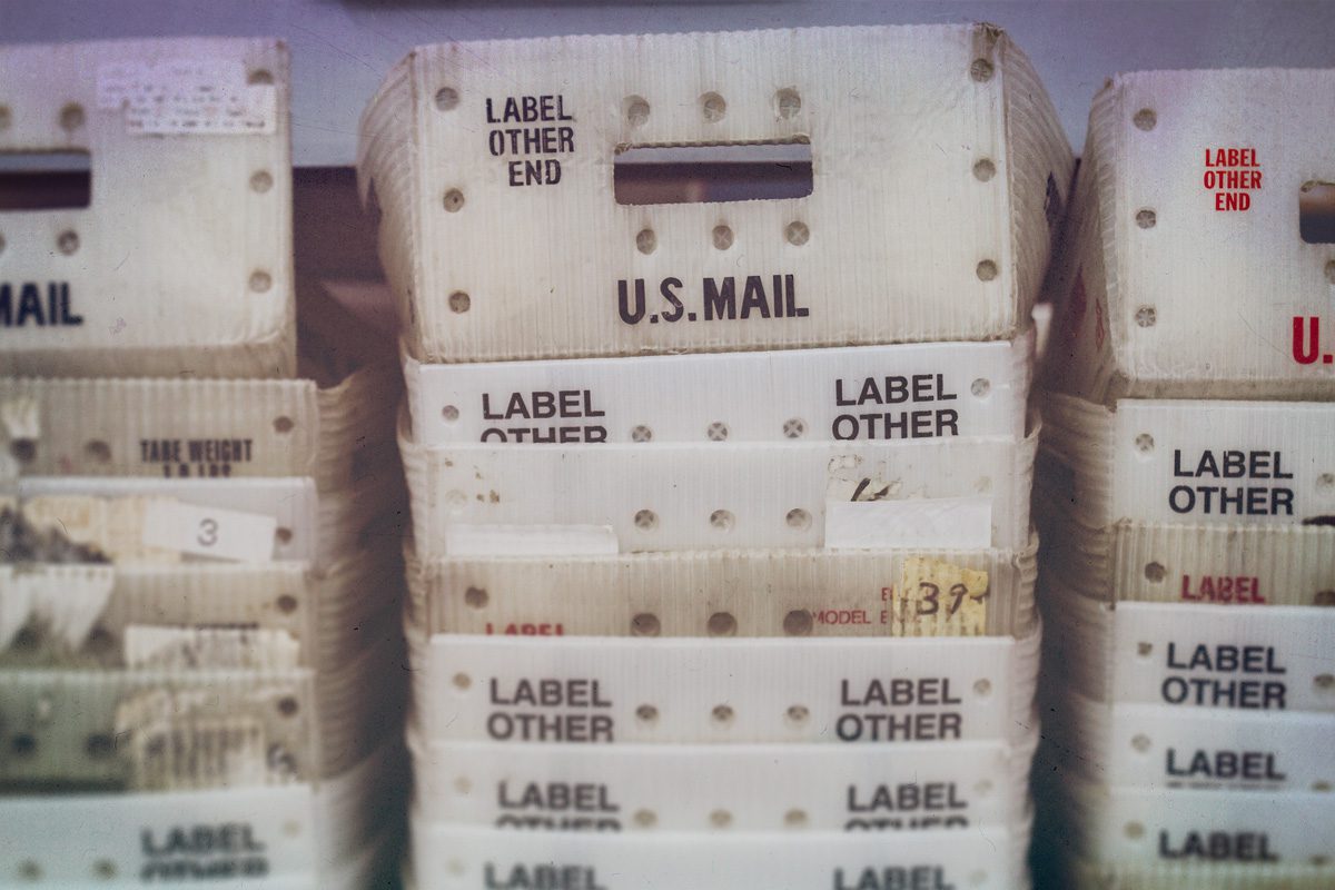 Stacked white plastic bins labeled U.S. Mail and Label Other End, with visible slots and wear marks, used for sorting and organizing mail.