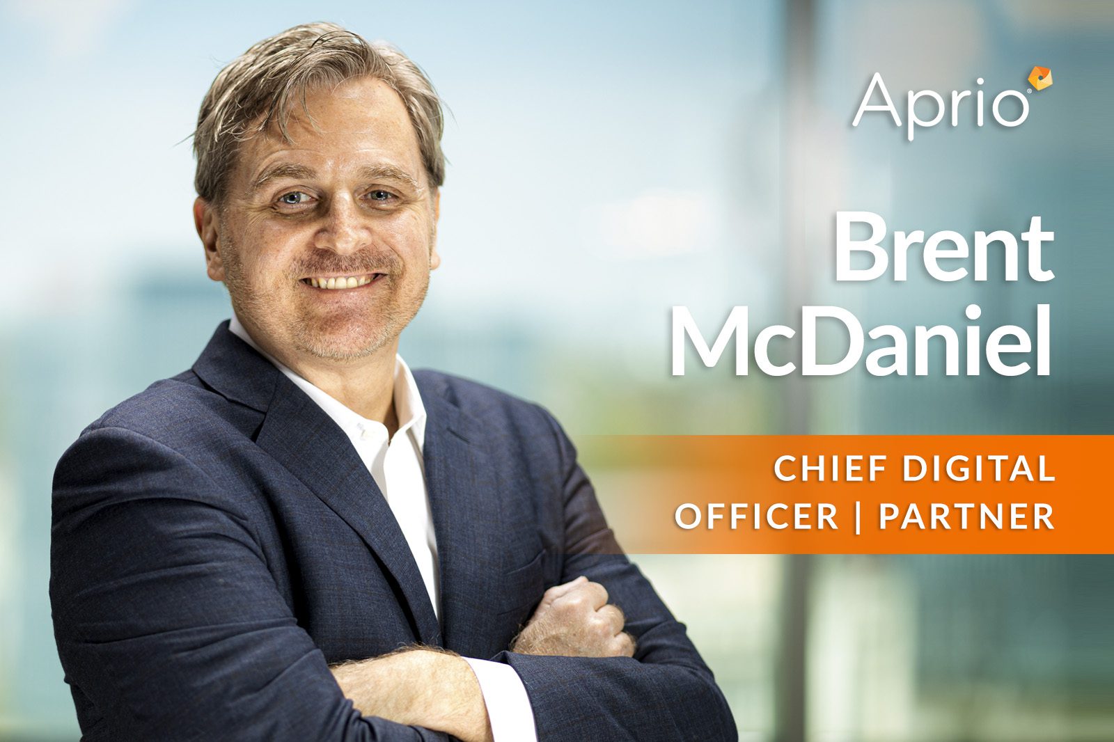 A man in a suit stands smiling with arms crossed. Text reads: Aprio. Brent McDaniel. Chief Digital Officer | Partner. A cityscape is visible in the blurred background.