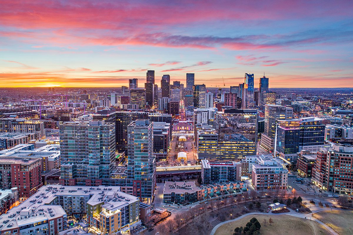 Aerial view of a modern city skyline at sunset, with tall buildings, residential areas, and a park in the foreground under a colorful sky.