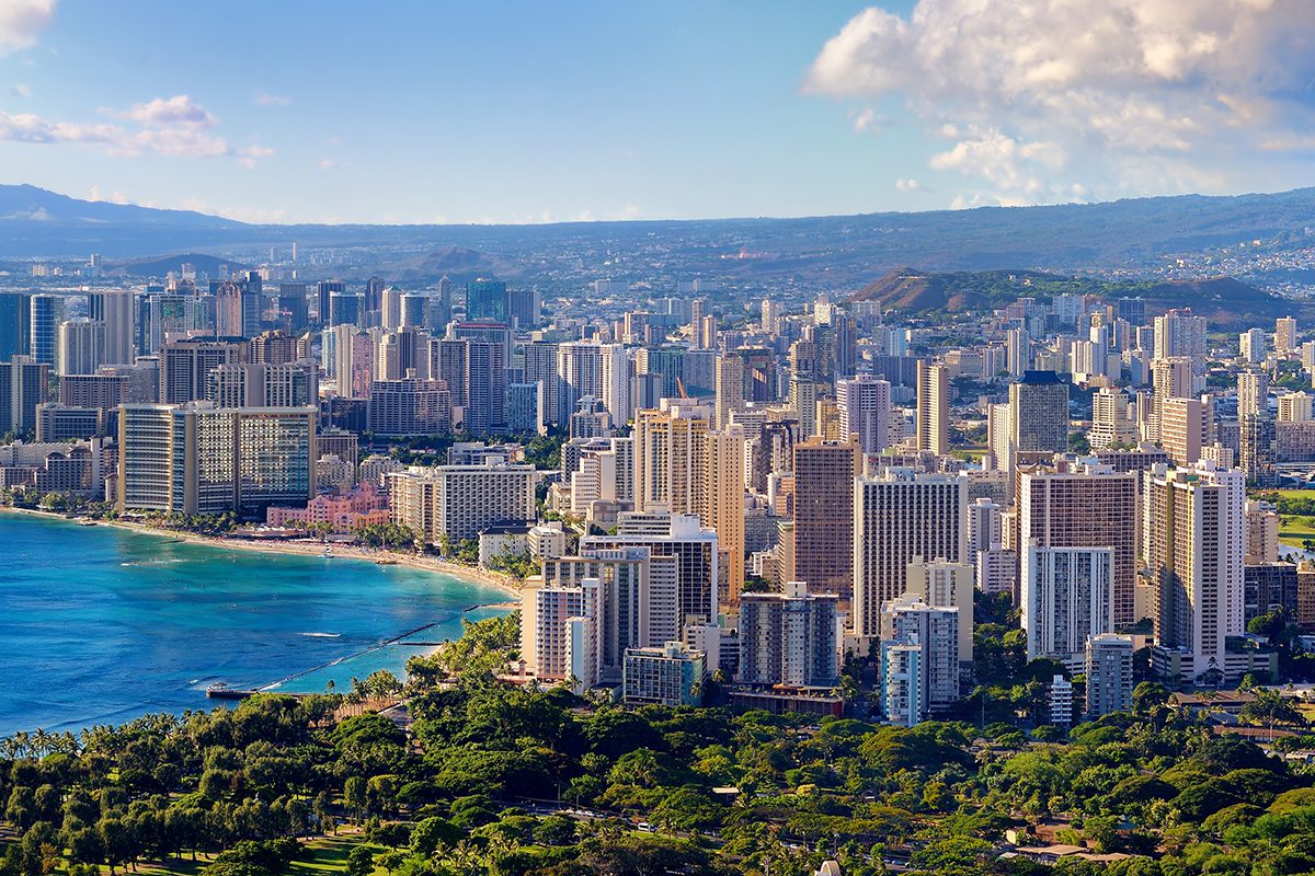 Aerial view of a coastal city with high-rise buildings, a curved shoreline, blue ocean water, and green park areas under a partly cloudy sky.