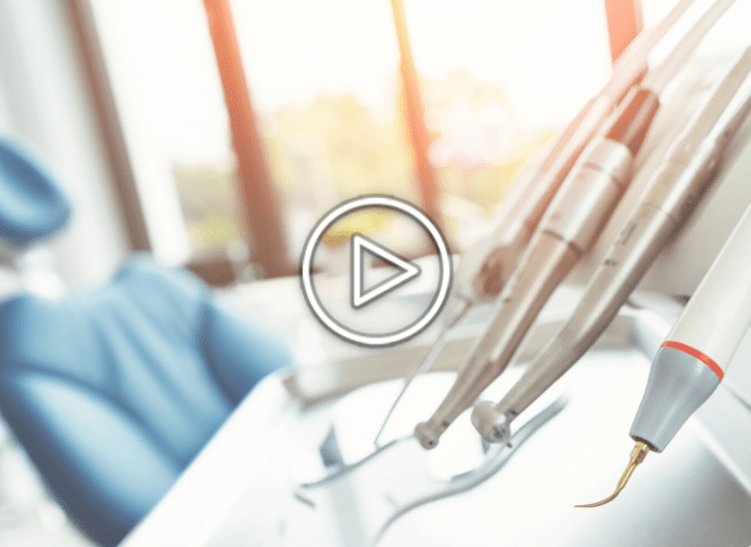 Dental tools on a tray in a bright clinic with a blue dental chair in the background; a play button icon is overlaid on the image.