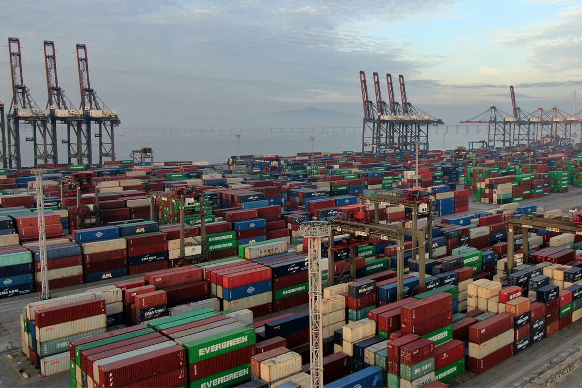 A large shipping port with numerous stacked cargo containers and several tall cranes near the waterfront under a cloudy sky.