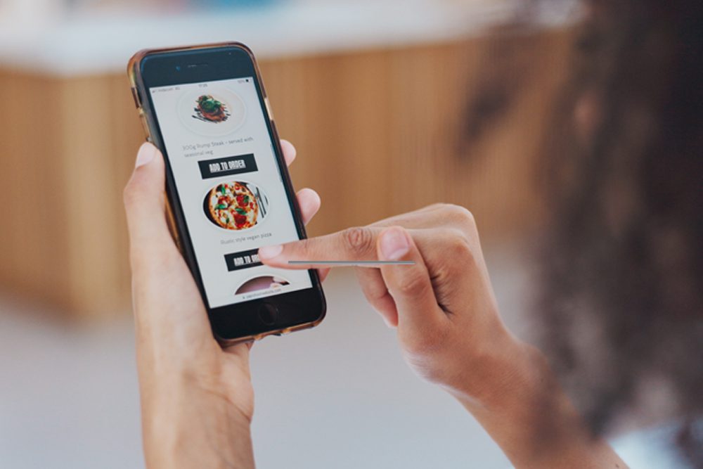 A person uses a smartphone to order food online, tapping a button under a pizza photo on a mobile app interface.