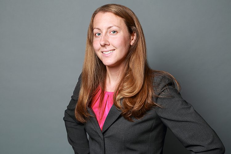 Woman with long brown hair wearing a dark gray blazer and pink top, standing in front of a plain gray background, smiling at the camera.