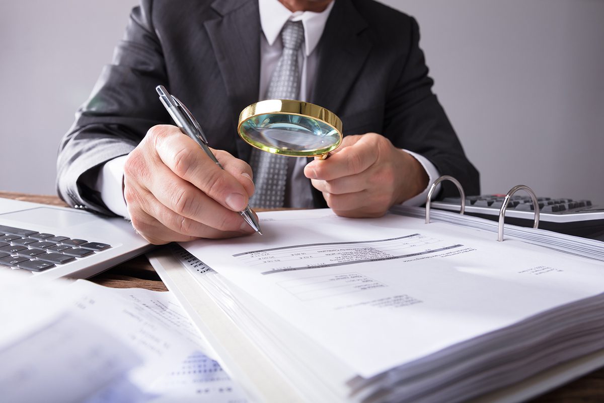 A person in a suit examines financial documents with a magnifying glass and pen at a desk with a laptop and binder.