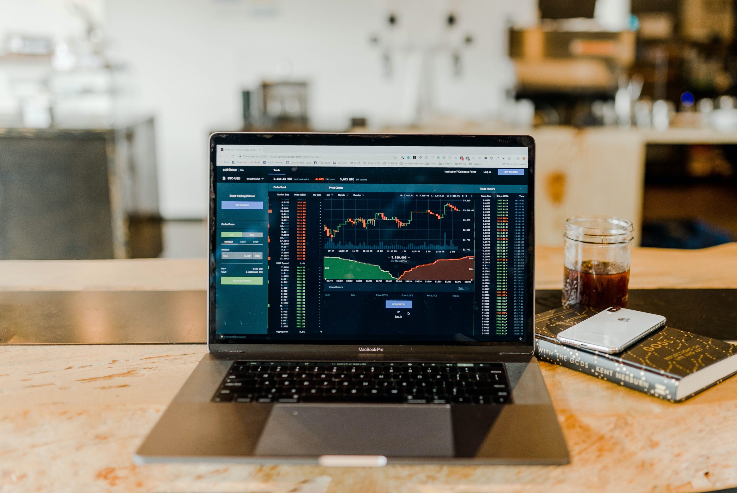 A laptop displaying a financial trading chart sits on a wooden table next to a glass of iced drink, a notebook, and a smartphone in a bright indoor setting.