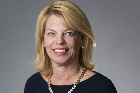 A woman with blonde hair, wearing a navy blue top and a gold chain necklace, smiles at the camera against a plain gray background.