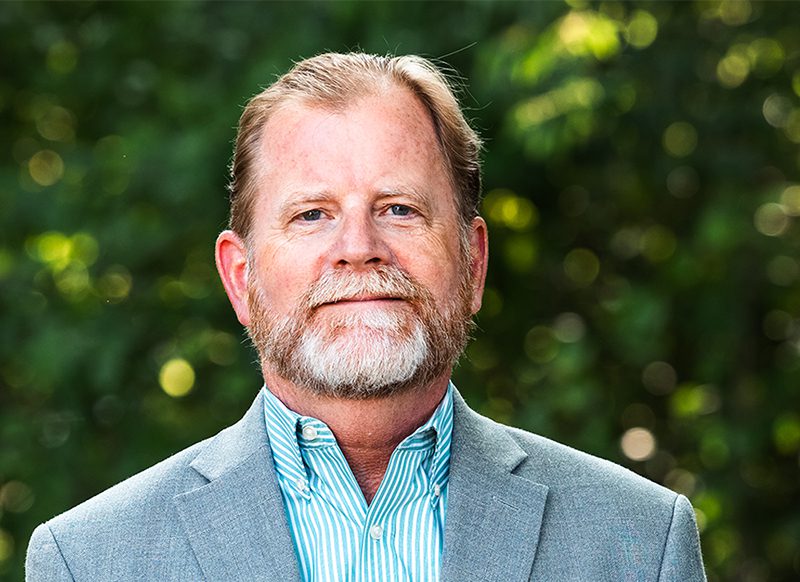 A middle-aged man with a beard and short hair wearing a light gray blazer and striped shirt stands outdoors in front of blurred greenery.