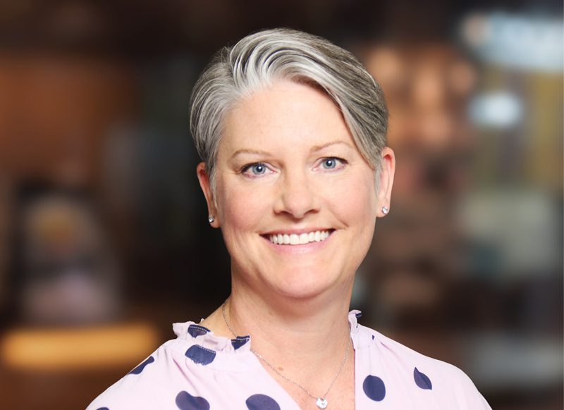 A woman with short, gray and brown hair smiles at the camera. She is wearing a light pink blouse with navy polka dots and small earrings. The background is softly blurred.