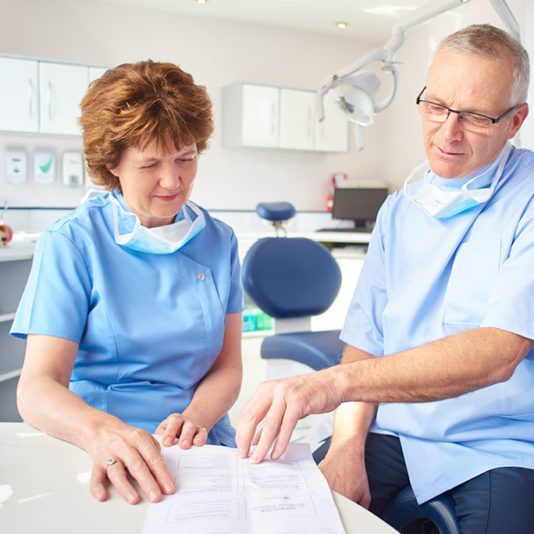 Two dental professionals wearing scrubs and face masks review documents together in a dental office.