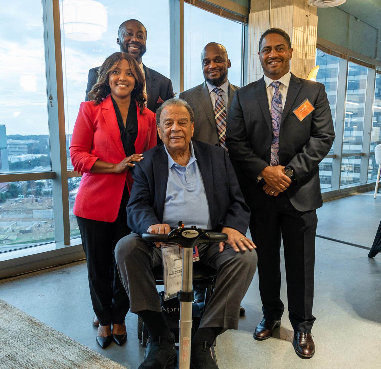 Five professionally dressed people pose indoors by large windows; one man is seated on a mobility scooter while the others stand behind him, smiling at the camera.