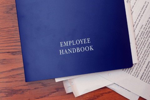 A blue booklet labeled Employee Handbook sits on top of several printed documents on a wooden surface.