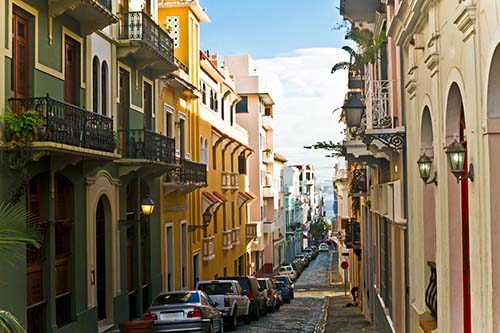 A narrow cobblestone street lined with parked cars and colorful colonial buildings with balconies on both sides, under a partly cloudy sky.