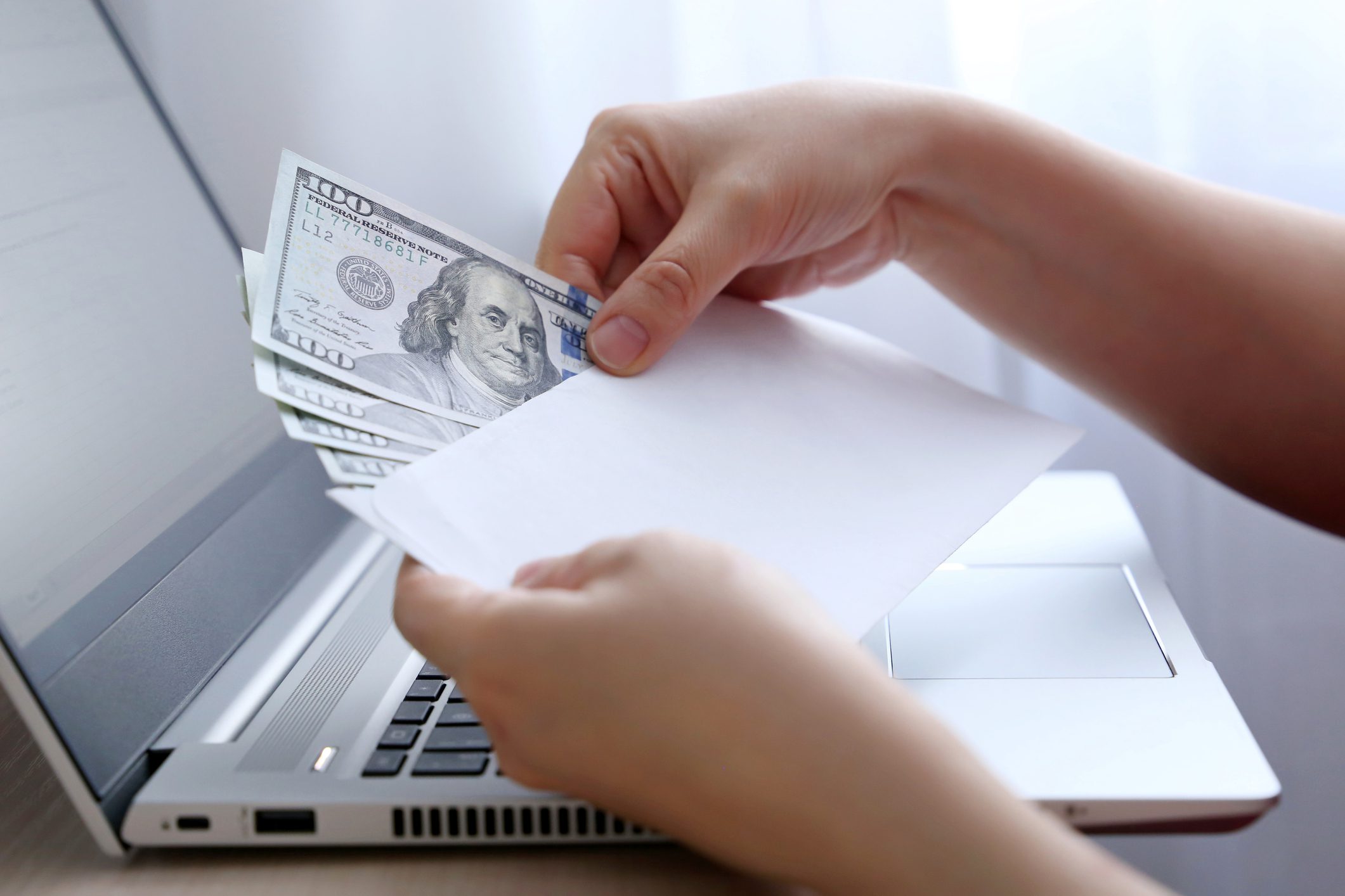 A person places several $100 bills into a white envelope in front of an open laptop.