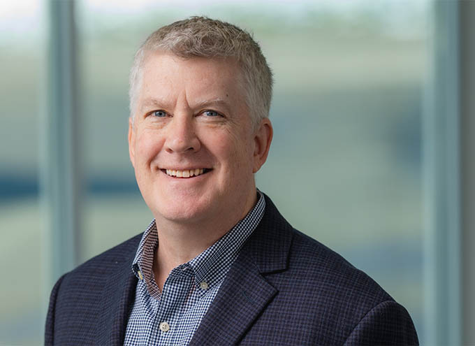 A middle-aged man with short gray hair wearing a dark plaid blazer and checkered shirt, smiling in front of a blurred window background.