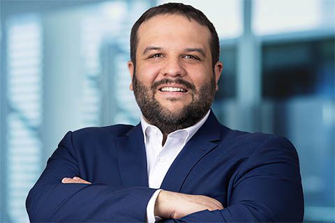 A man with a beard and short hair, wearing a navy blue suit and white shirt, stands with arms crossed and smiles at the camera in an office setting.