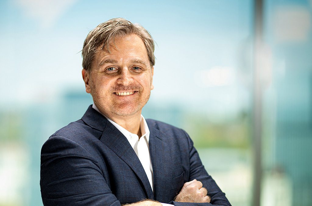 A man in a blue suit and white shirt stands with arms crossed, smiling at the camera, with a blurred office background.