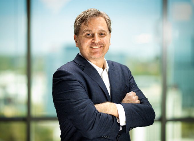 A man in a dark suit with a white shirt stands indoors with his arms crossed, smiling at the camera. Blurred cityscape seen through large windows in the background.