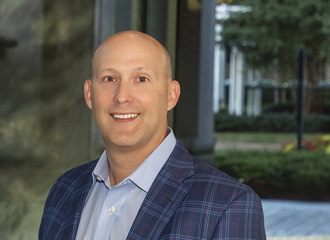 A man in a blue plaid suit jacket and light blue shirt smiles at the camera outdoors, with greenery and buildings blurred in the background.