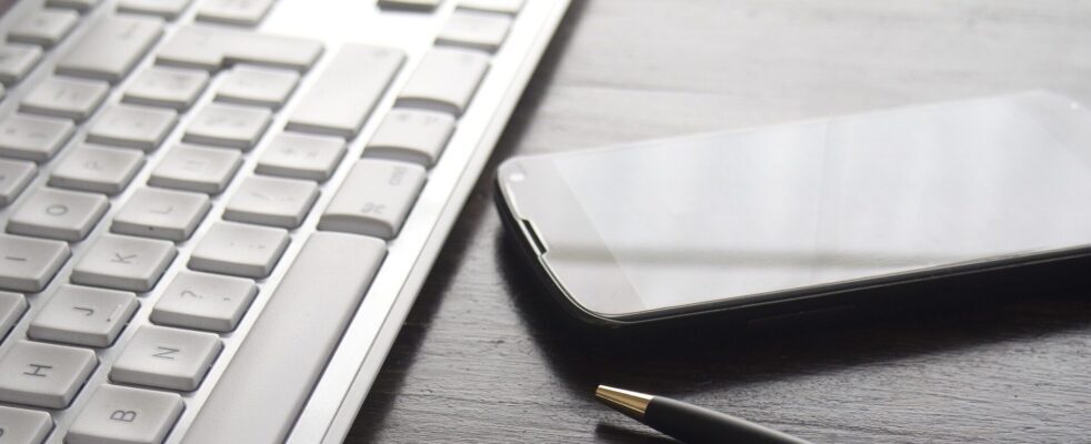 A close-up of a white computer keyboard, a black smartphone, and a pen on a dark wooden desk.