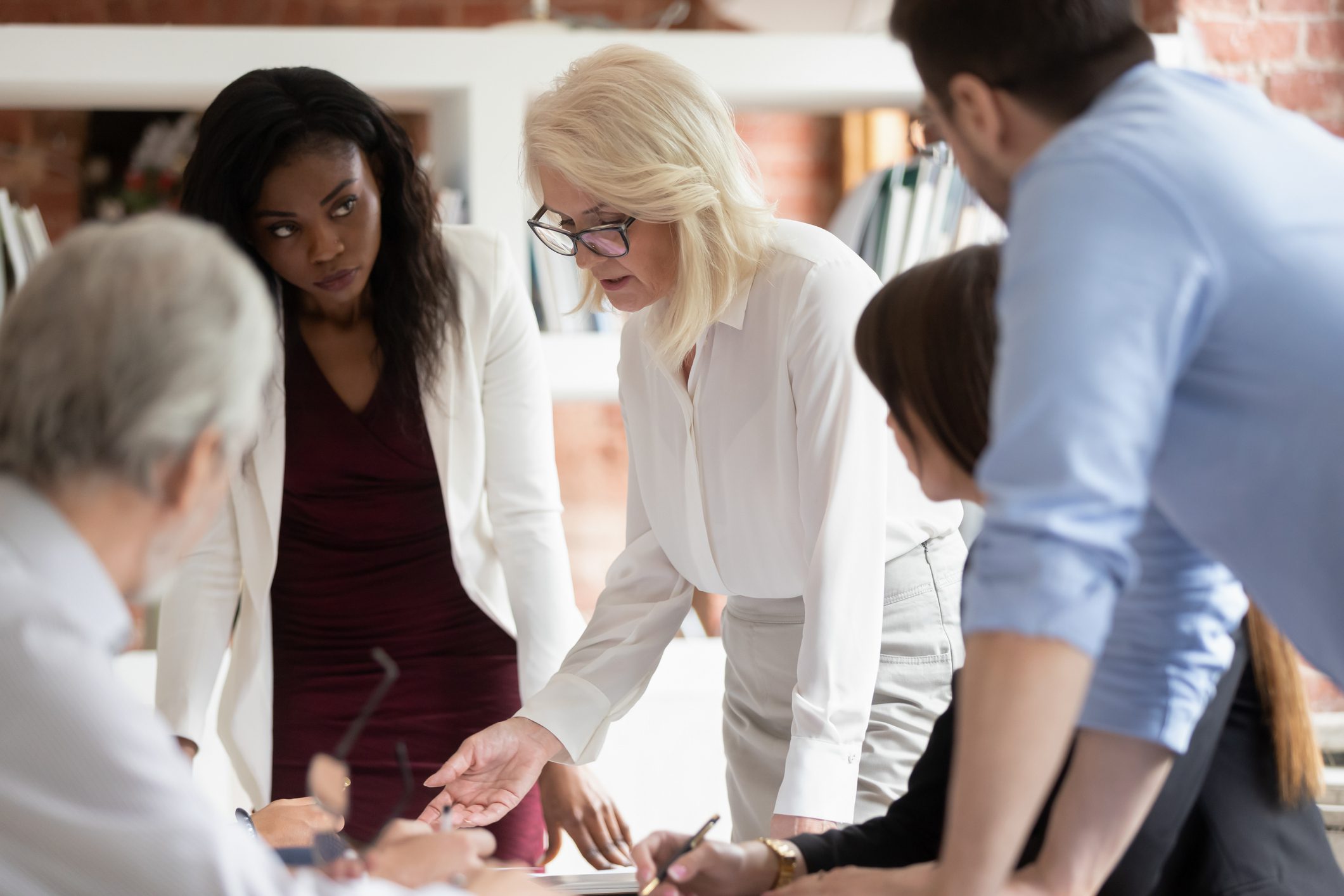 A group of six professionals gathered around a table in discussion, with one woman standing and speaking while others listen and take notes.