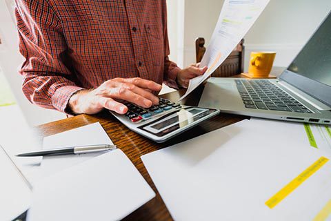 Person using a calculator while holding a document, with a laptop, pen, and papers on a wooden desk.