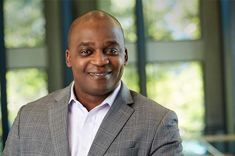 A man in a gray checked suit jacket and light shirt smiles at the camera in an indoor setting with large windows and greenery outside.