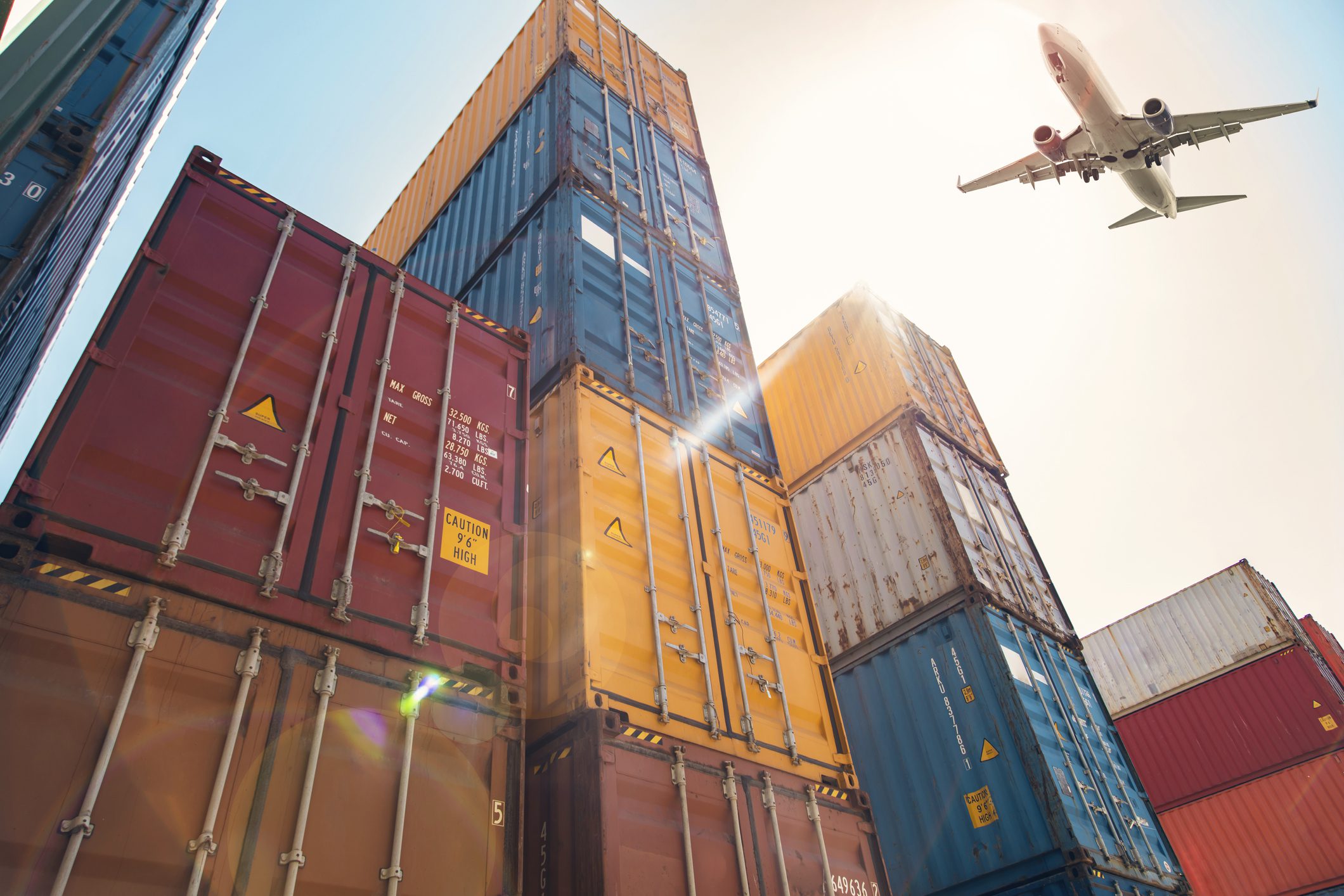 Tall stacks of colorful shipping containers under bright sunlight, with a commercial airplane flying overhead in a clear sky.