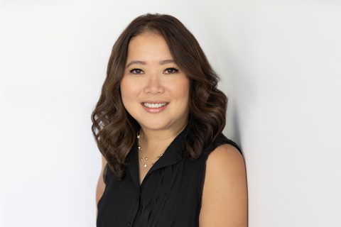 Woman with medium-length wavy brown hair, wearing a sleeveless black top, smiles in front of a plain white background.