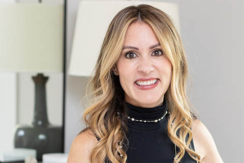 A woman with long, wavy blonde hair, wearing a sleeveless black top, smiles at the camera. A lamp and mirror are visible in the background.