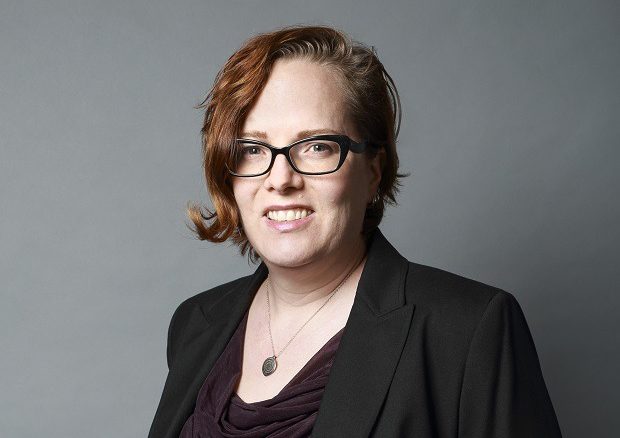 Person with short, light brown hair and glasses, wearing a black blazer and necklace, stands against a plain grey background, smiling slightly at the camera.