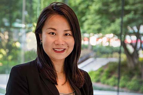 A woman with straight dark hair, wearing a black blazer and gray top, stands indoors in front of a window with greenery outside.