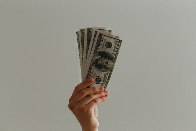 A hand holds up several U.S. hundred dollar bills against a plain, light background.