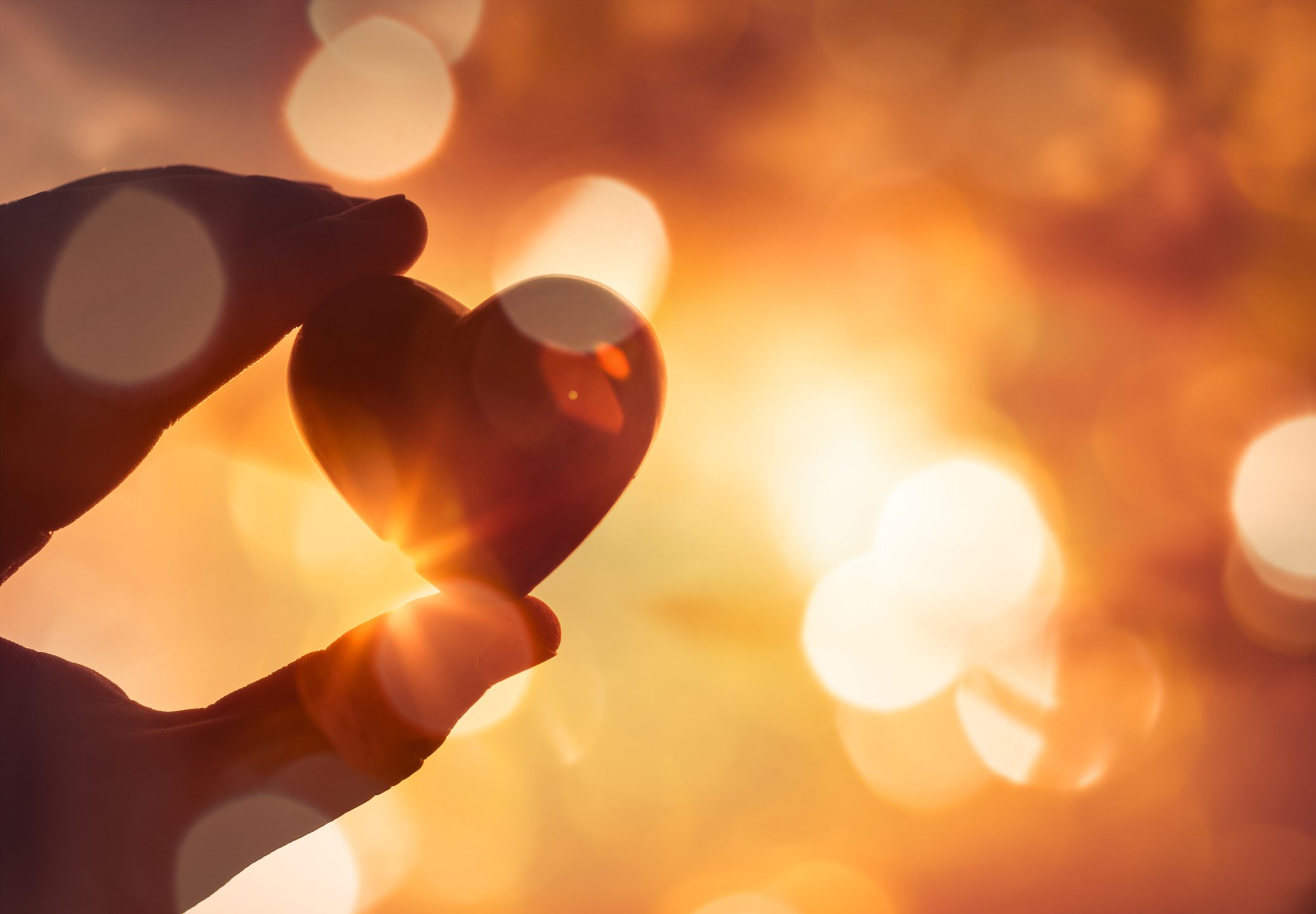 A hand holds a small heart-shaped object against a bright, golden, and softly blurred background with bokeh light effects.