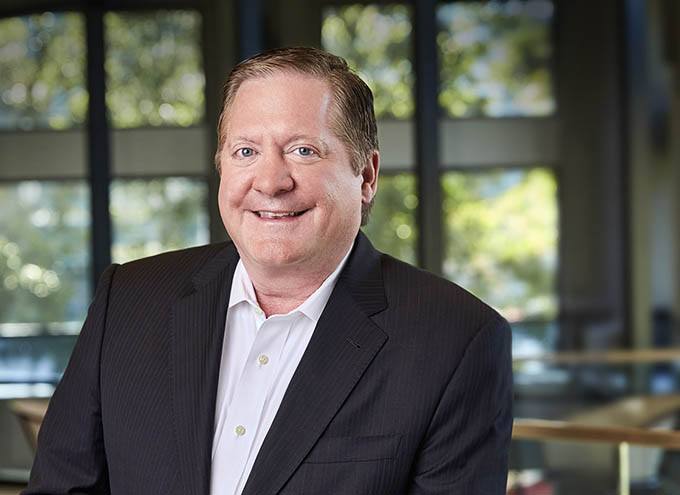 A middle-aged man in a dark suit and white shirt is smiling, standing indoors with blurred windows and trees in the background.