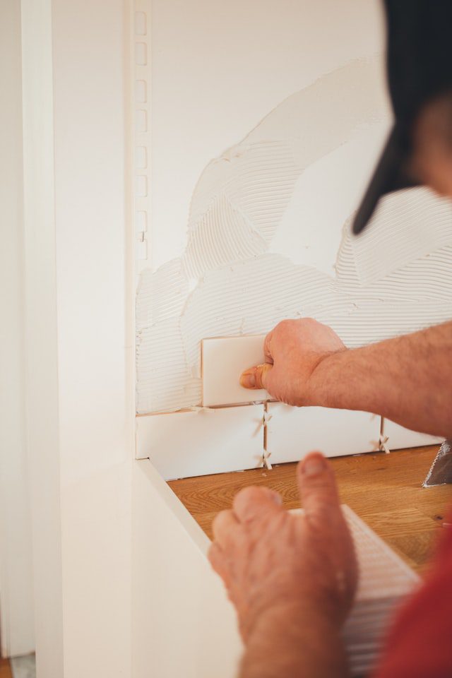 A person installs white ceramic tiles on a wall, using a trowel to spread adhesive and align the tiles.