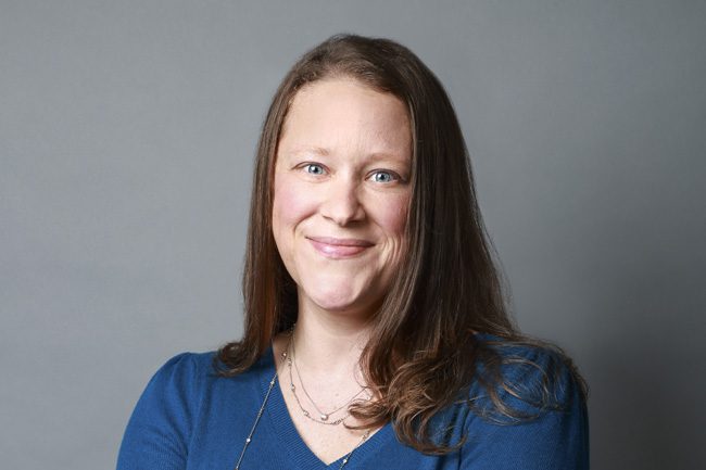 A woman with long brown hair, wearing a blue top and necklaces, smiles at the camera against a plain gray background.