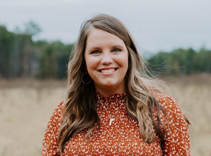 A woman with long brown hair wearing a rust-colored patterned blouse stands outdoors in a grassy field, smiling at the camera.