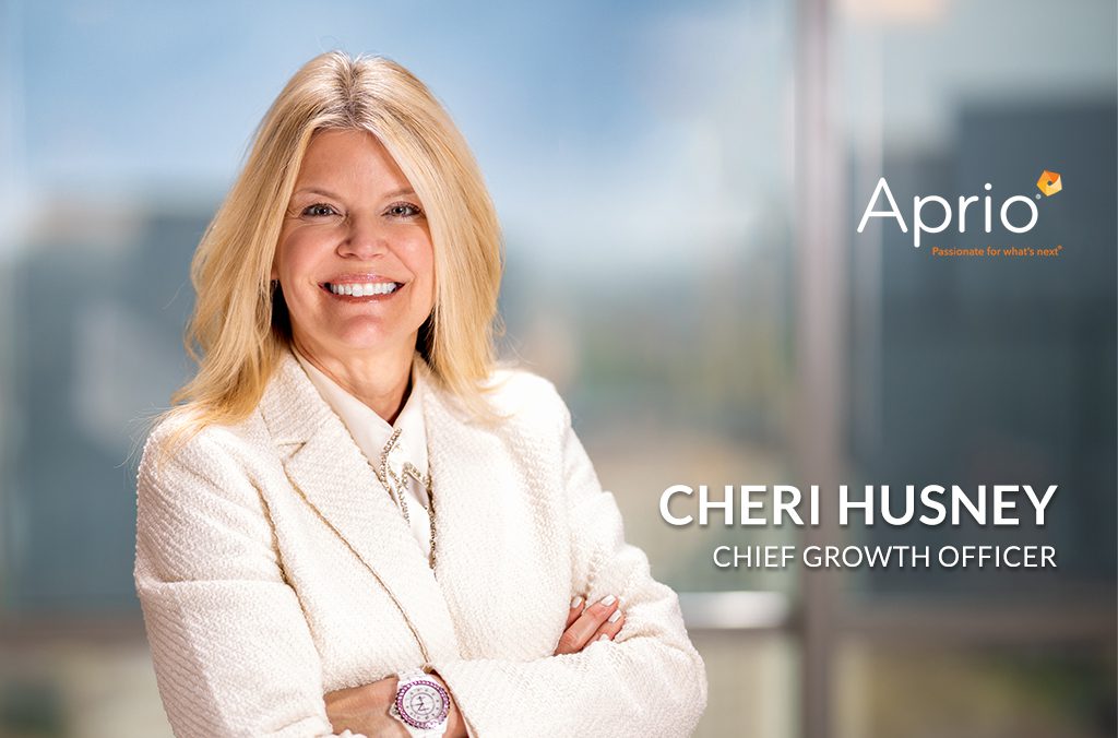 A woman in business attire stands smiling with arms crossed. Text on image: Aprio, Cheri Husney, Chief Growth Officer. A blurred office background is visible.