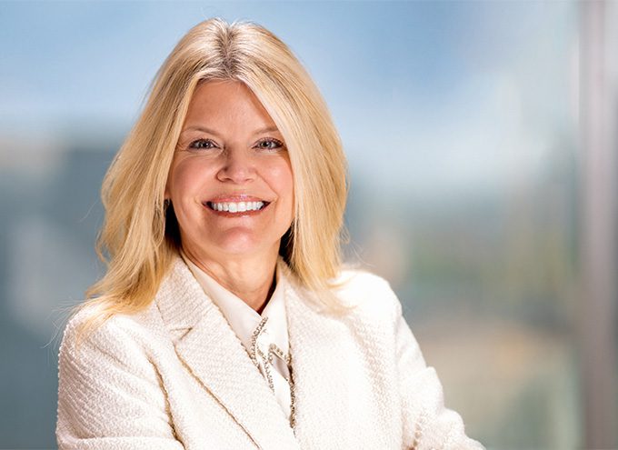 A woman with long blonde hair, wearing a white blazer and blouse, smiles at the camera against a blurred background.